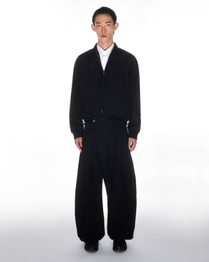 A person stands facing forward against a white background, wearing a black zip-up jacket, a white collared shirt, Sundae School's BLACK PLEATED NYLON PANTS, and black shoes.