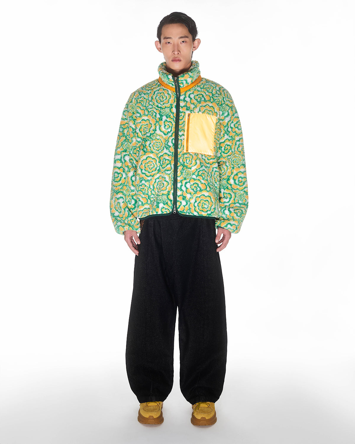 A person faces forward against a white background, wearing Sundae School's Broccoli Fleece—a green and yellow puffer with a custom zipper and yellow chest pocket—paired with black wide-leg pants and yellow shoes.