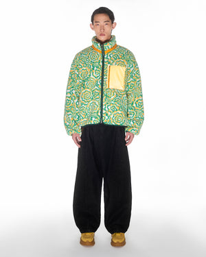 A person faces forward against a white background, wearing Sundae School's Broccoli Fleece—a green and yellow puffer with a custom zipper and yellow chest pocket—paired with black wide-leg pants and yellow shoes.