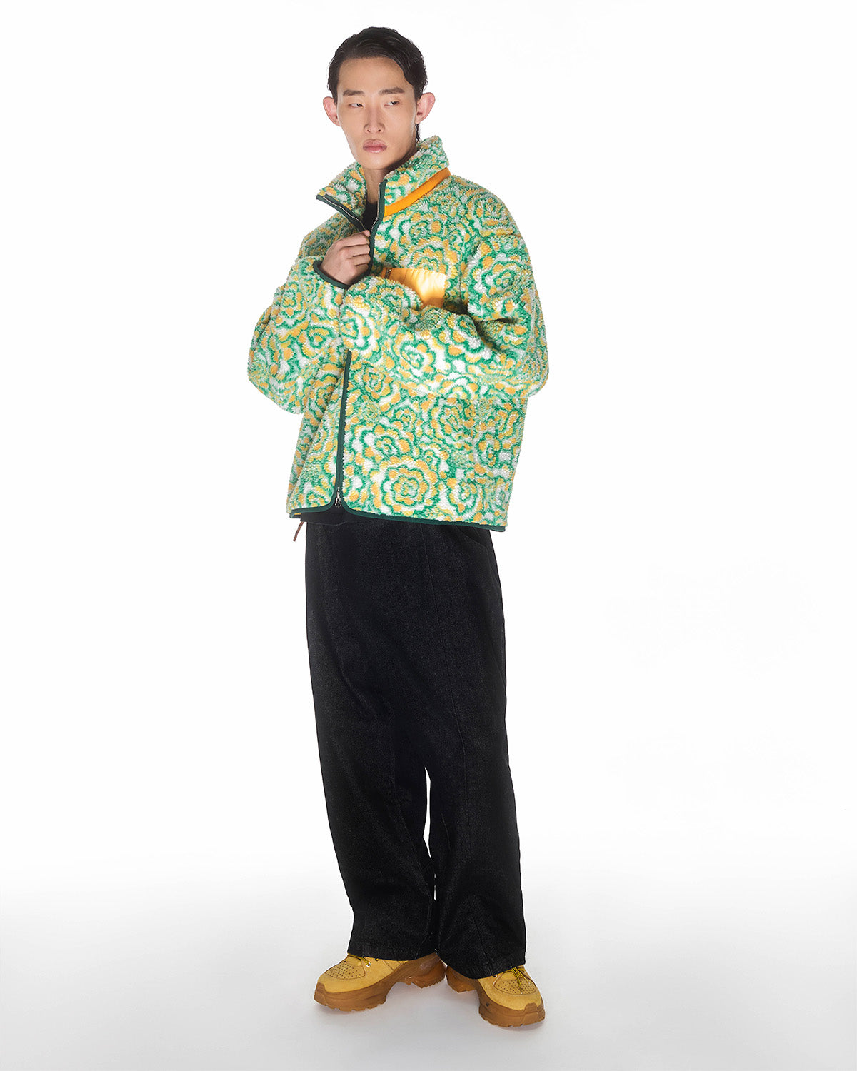 A person with short dark hair stands against a white background, looking at the camera while holding the collar of the Sundae School Broccoli Fleece jacket, paired with black wide-leg pants and yellow shoes.