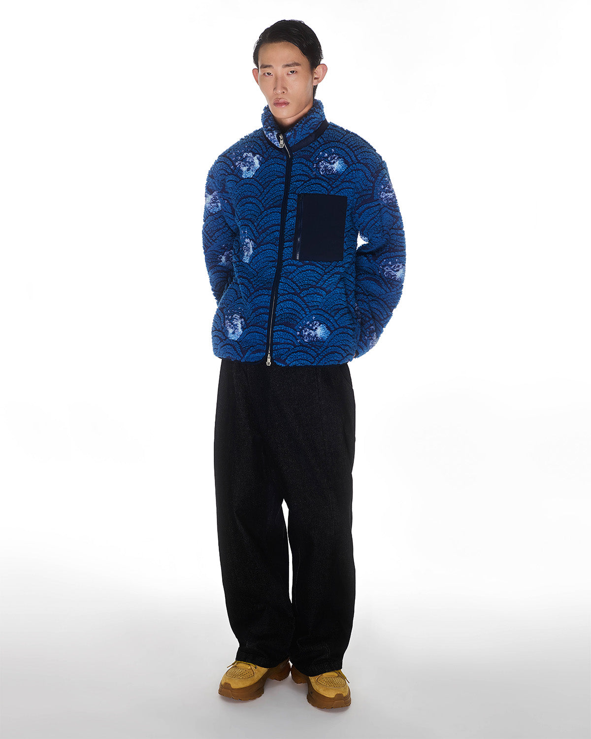 A person stands against a white background wearing the Sundae School BLUE WAVE FLEECE, featuring an embossed wave motif, paired with loose black pants and tan shoes. Their hands are in their pockets and they have a neutral expression.