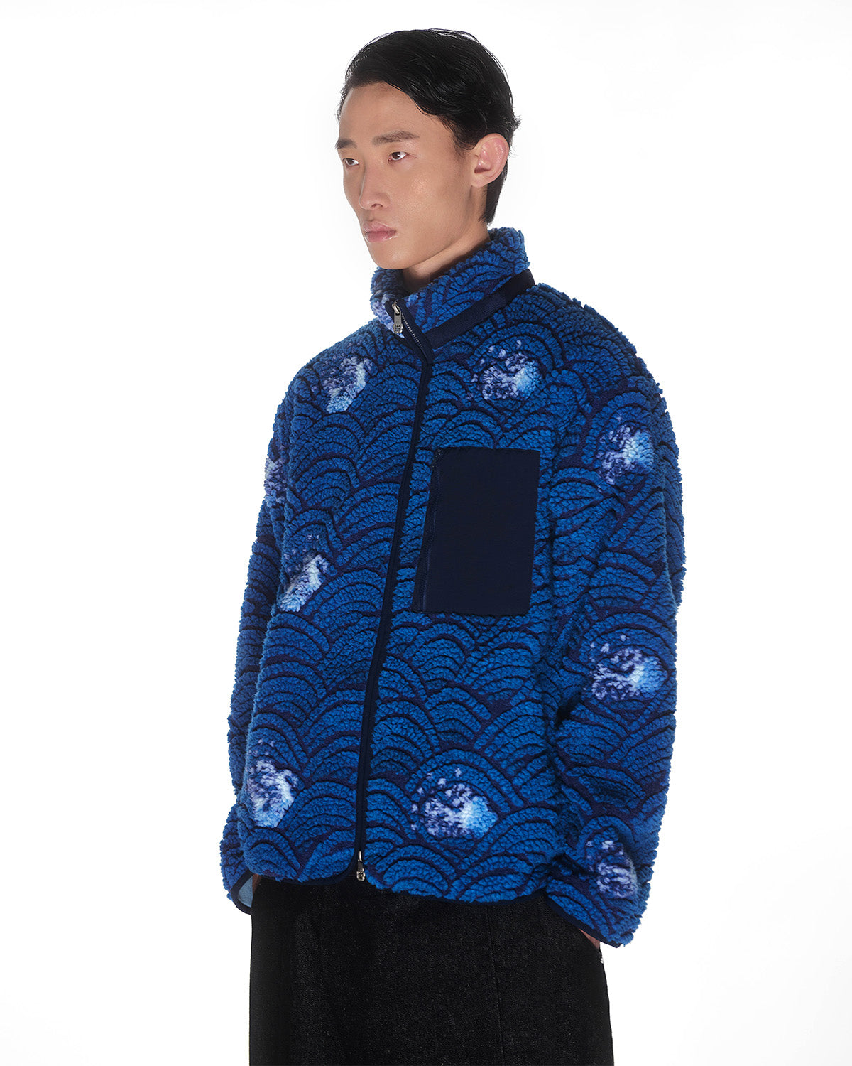 A person wears Sundae School’s BLUE WAVE FLEECE, featuring a custom wave motif jacquard, standing against a plain white background with hands in pockets and looking slightly to the side.