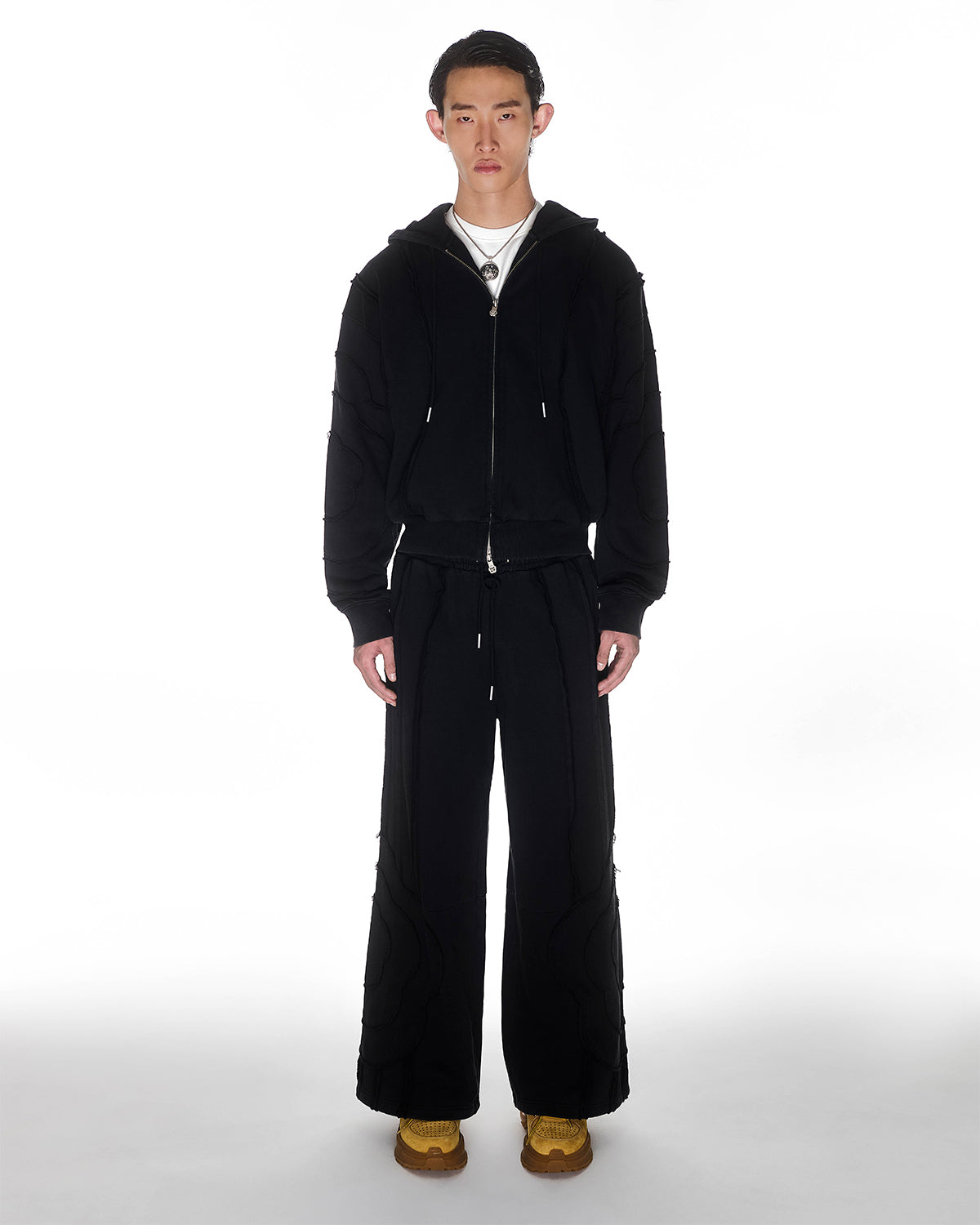 Black Mochi Haze Paneled Hoodie featuring black garment washed cotton with exposed curved seam details, paired with matching black Mochi Haze Paneled Sweatpants, zip-up construction with drawstring hood, relaxed oversized fit silhouette, modeled on light background, shown in front view.