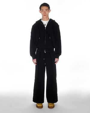 Black Mochi Haze Paneled Hoodie featuring black garment washed cotton with exposed curved seam details, paired with matching black Mochi Haze Paneled Sweatpants, zip-up construction with drawstring hood, relaxed oversized fit silhouette, modeled on light background, shown in front view.