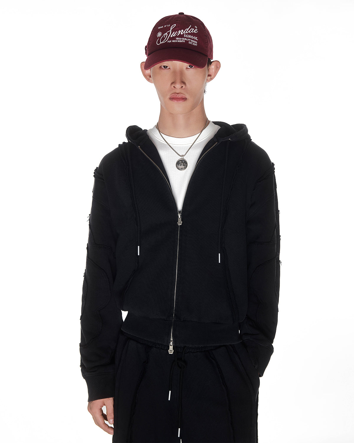 Black Mochi Haze Paneled Hoodie featuring garment washed black cotton with exposed curved seam details, paired with matching black Mochi Haze Paneled Sweatpants, styled with burgundy baseball cap and silver chain necklace, relaxed oversized fit silhouette, modeled on light background, shown in front view.