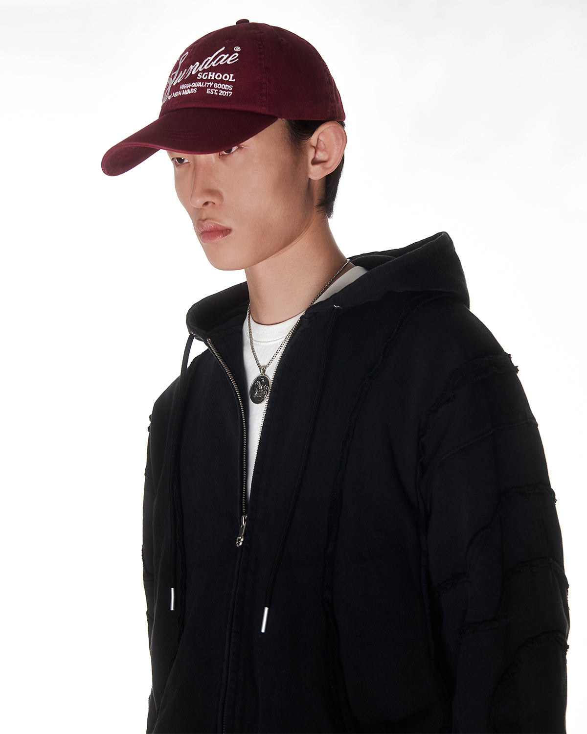 Black Mochi Haze Paneled Hoodie styled with burgundy baseball cap and silver chain necklace, cropped to show upper chest and face area, zip-up construction with drawstring hood, garment washed black cotton with exposed seam details, photographed on light background.