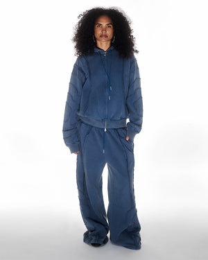 Indigo Mochi Haze Paneled Hoodie featuring garment washed indigo cotton with exposed curved seam details paired with matching indigo Mochi Haze Paneled Sweatpants, zip-up construction with drawstring hood, relaxed oversized fit silhouette, modeled on light background, shown in front view.