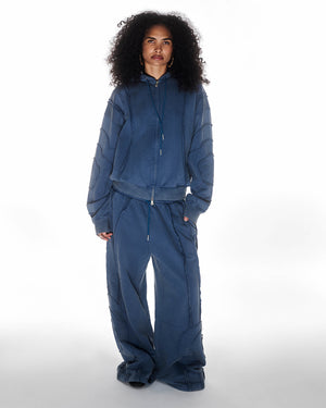 Indigo Mochi Haze Paneled Hoodie featuring garment washed indigo cotton with exposed curved seam details, paired with matching indigo Mochi Haze Paneled Sweatpants, zip-up construction with drawstring hood, relaxed oversized fit silhouette, modeled on light background, shown in front view.