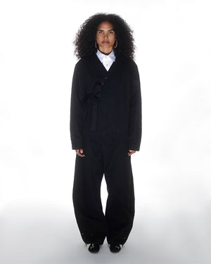 A person with curly hair stands against a white background, wearing the BLACK HANBOK TIE BLAZER by Sundae School with wide-legged pants, a white collared shirt, black shoes, gold hoop earrings, and a serious expression.