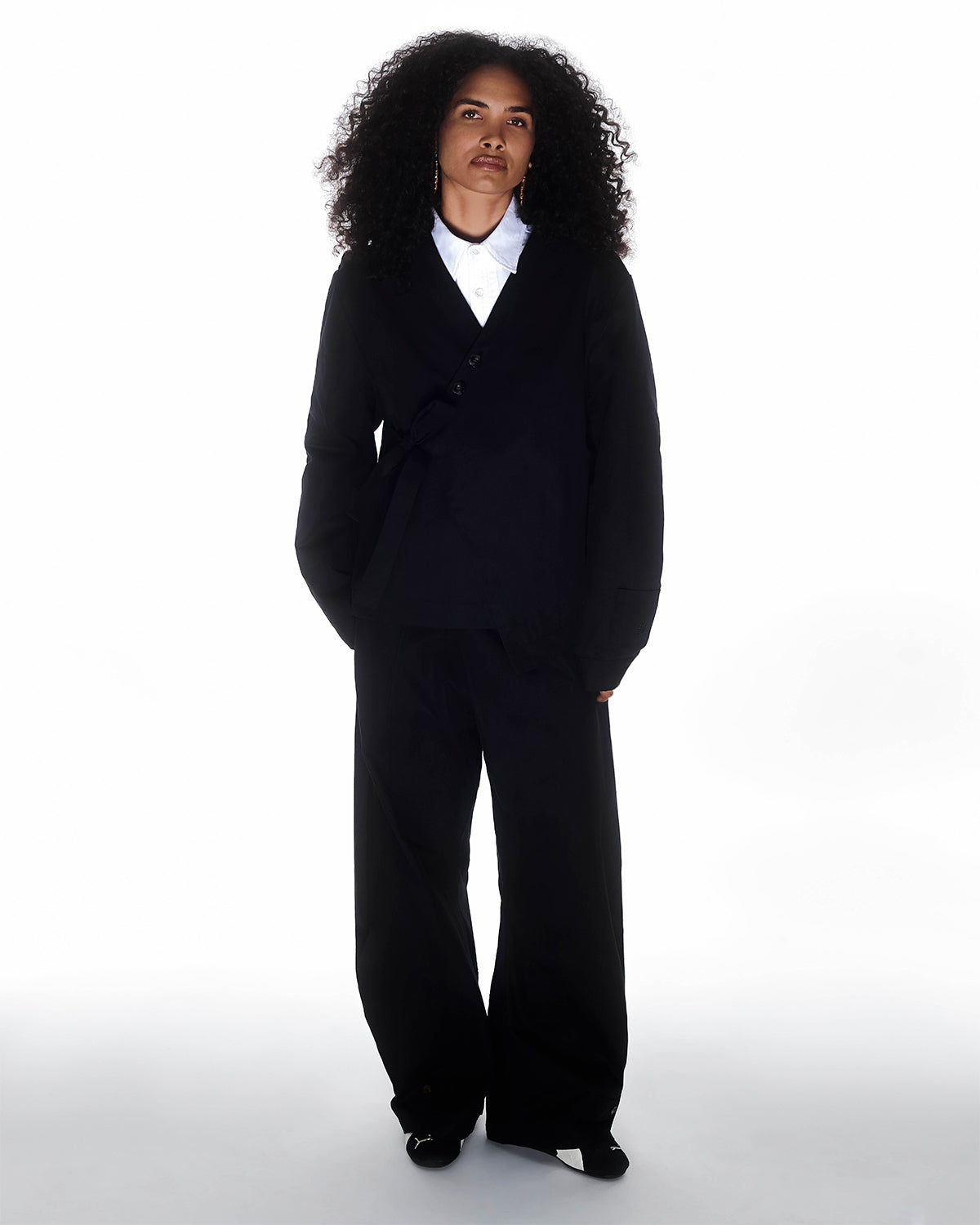 A person with curly hair stands against a white background, wearing Sundae School BLACK HANBOK BALLOON PANTS styled with a black wrap blazer and white collared shirt, hands in pockets and a neutral expression.