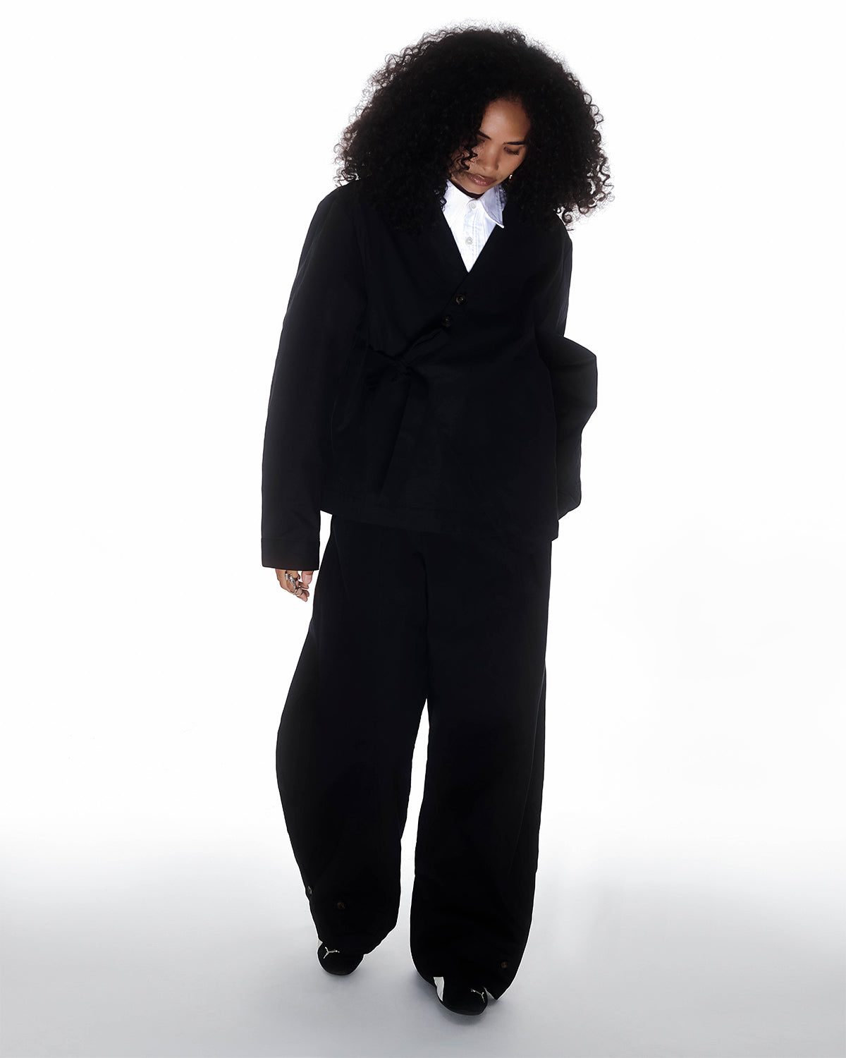 A person with curly hair wears the BLACK HANBOK TIE BLAZER by Sundae School—a loose black suit over a white collared shirt—standing against a white background and evoking a modern Hanbok silhouette.