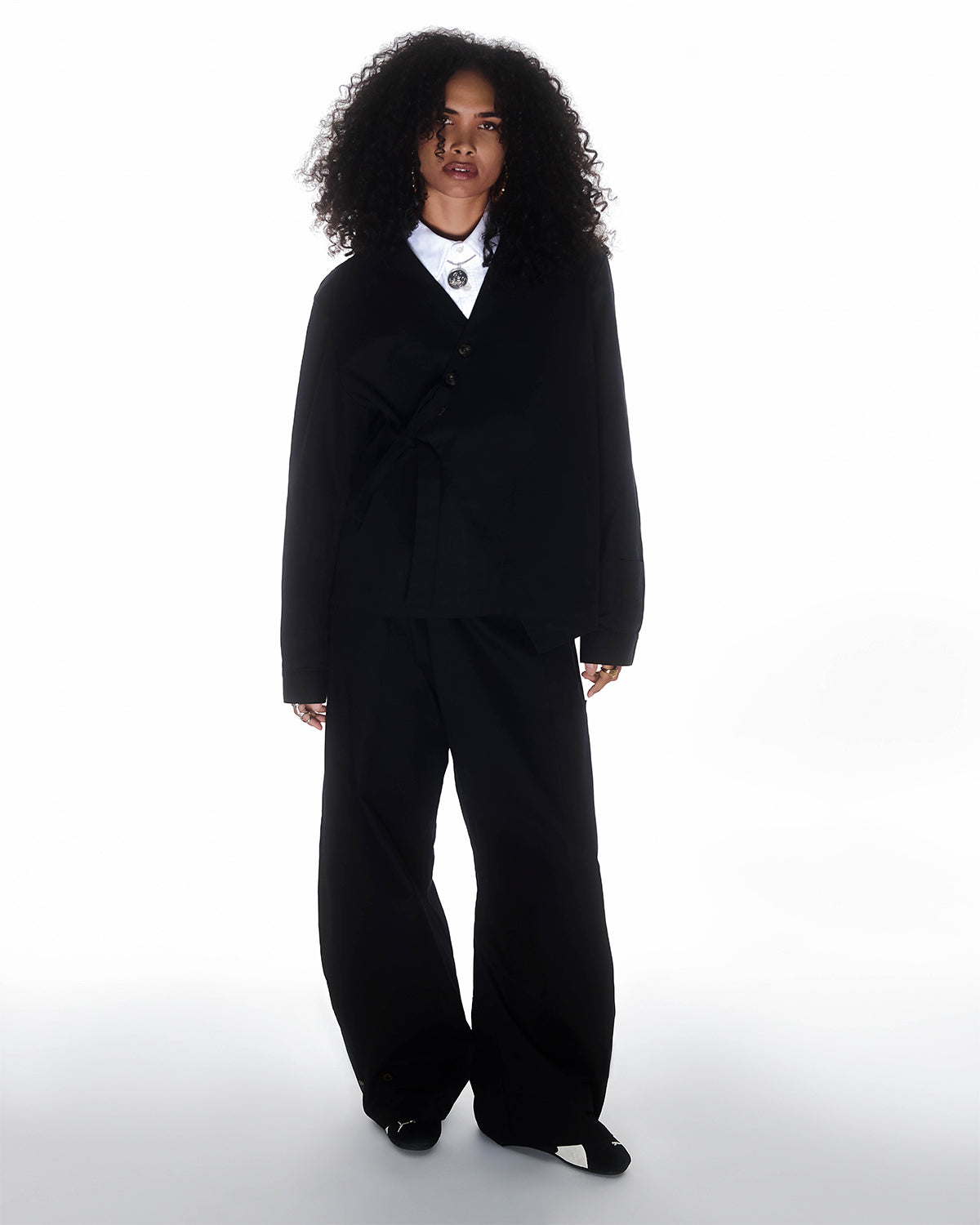 A woman with curly hair stands facing forward, wearing the BLACK HANBOK TIE BLAZER by Sundae School with wide-leg pants, a white collared shirt, and a pendant necklace against a plain white background.