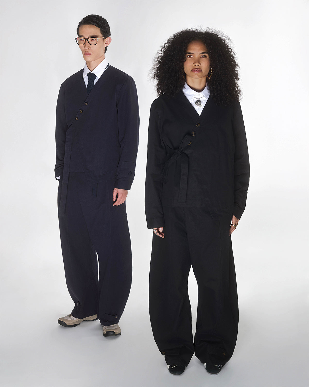 Two people stand side by side in a studio, wearing Sundae School's BLACK HANBOK TIE BLAZER over white collared shirts. One has curly hair, the other short straight hair and glasses. Both have neutral expressions.