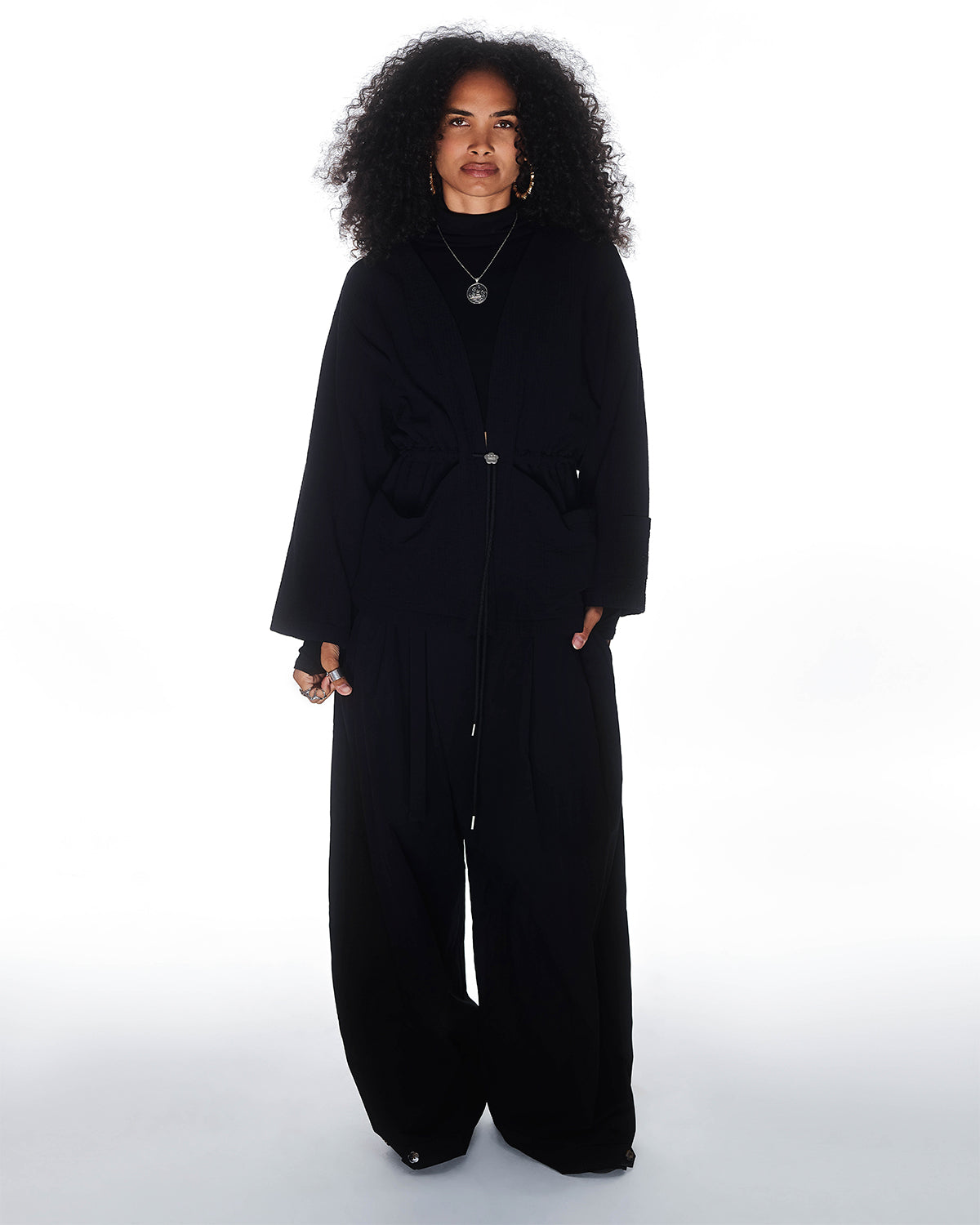 A person with curly hair wears wide-leg black pants, layered necklaces, hoop earrings, and the Sundae School QUILTED BOLO TIE CARDIGAN with a 떨 logo bolo tie, standing against a plain white background.