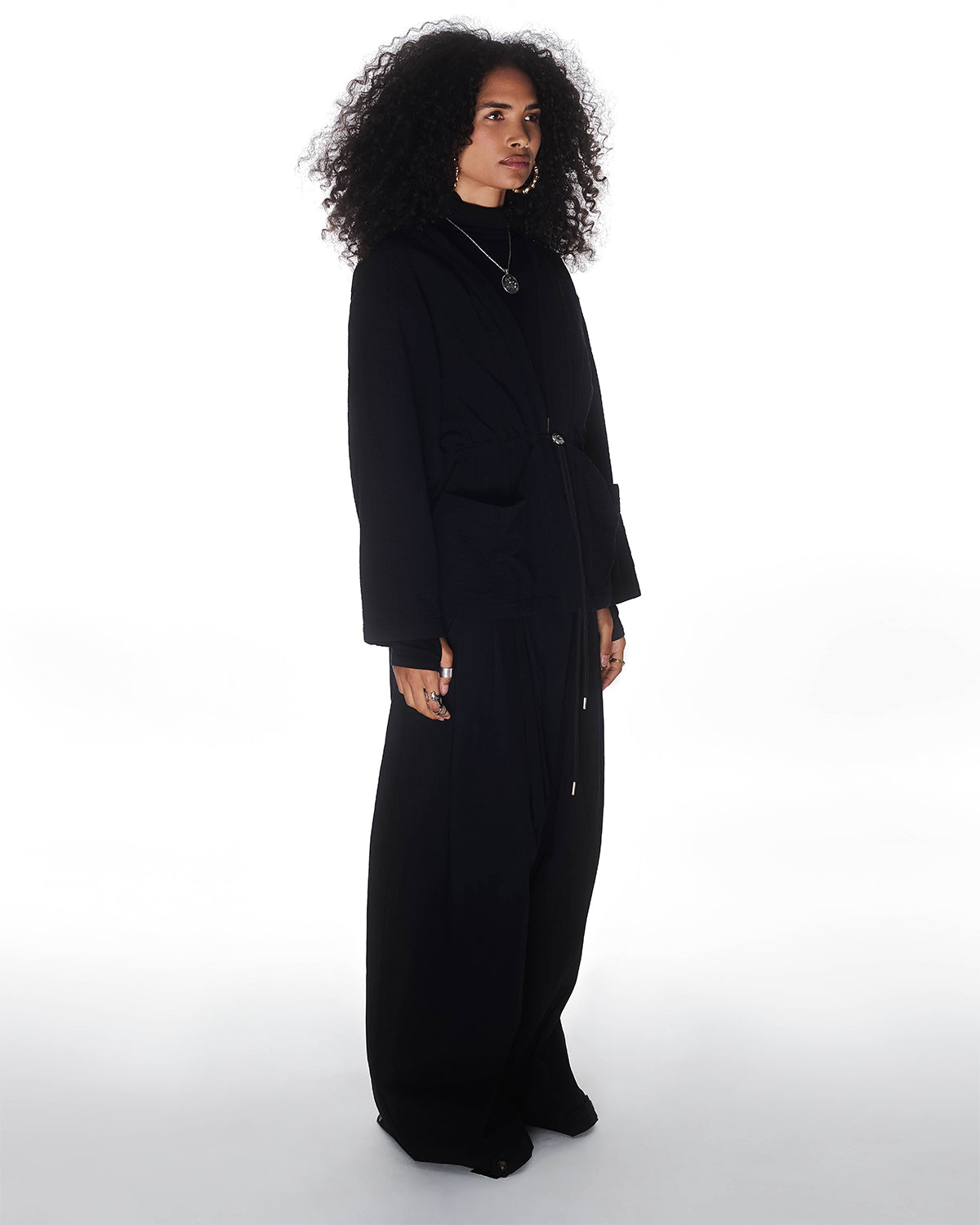 A person with curly hair stands against a white background, wearing Sundae School's QUILTED BOLO TIE CARDIGAN and wide-legged pants, completed with a 떨 logo bolo tie necklace.