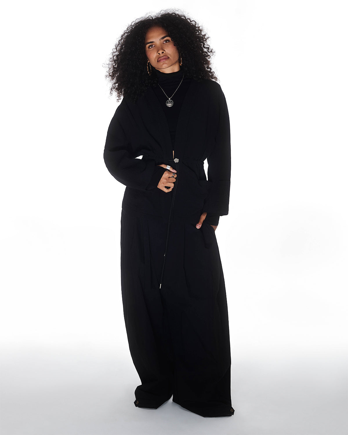 A person with curly hair, styled in the Sundae School QUILTED BOLO TIE CARDIGAN and layered necklaces, stands confidently against a white background, looking at the camera.