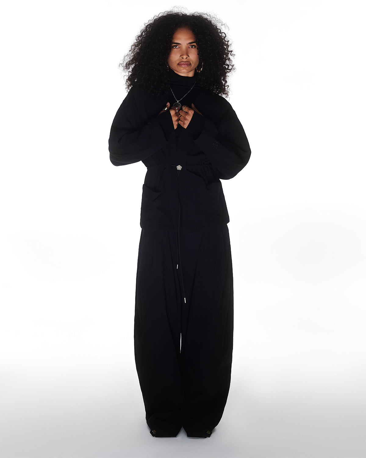 A person with curly hair stands facing forward against a white background, wearing the Sundae School QUILTED BOLO TIE CARDIGAN with wide-leg trousers and a buttoned-up jacket, hands held near their chest.