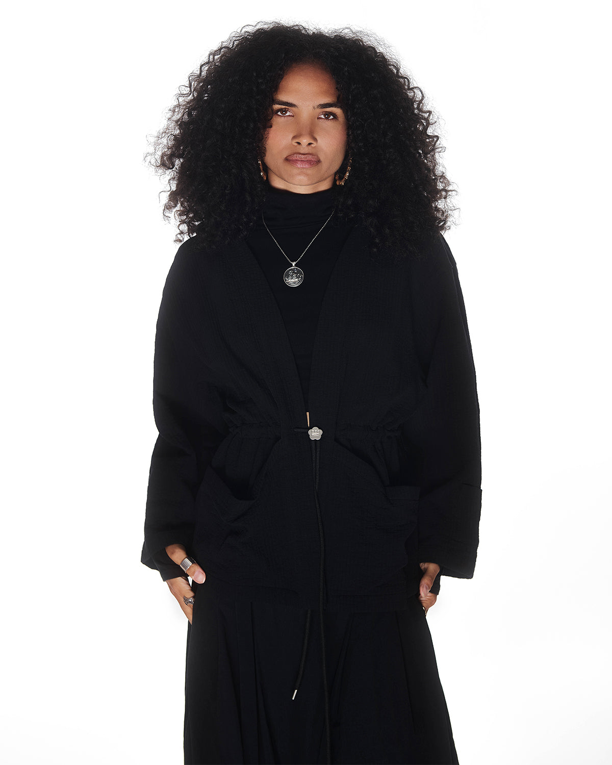 A person with long curly hair wears the Sundae School QUILTED BOLO TIE CARDIGAN over a black turtleneck and skirt, styled with layered necklaces, rings, and a 떨 logo bolo tie, standing against a plain white background.