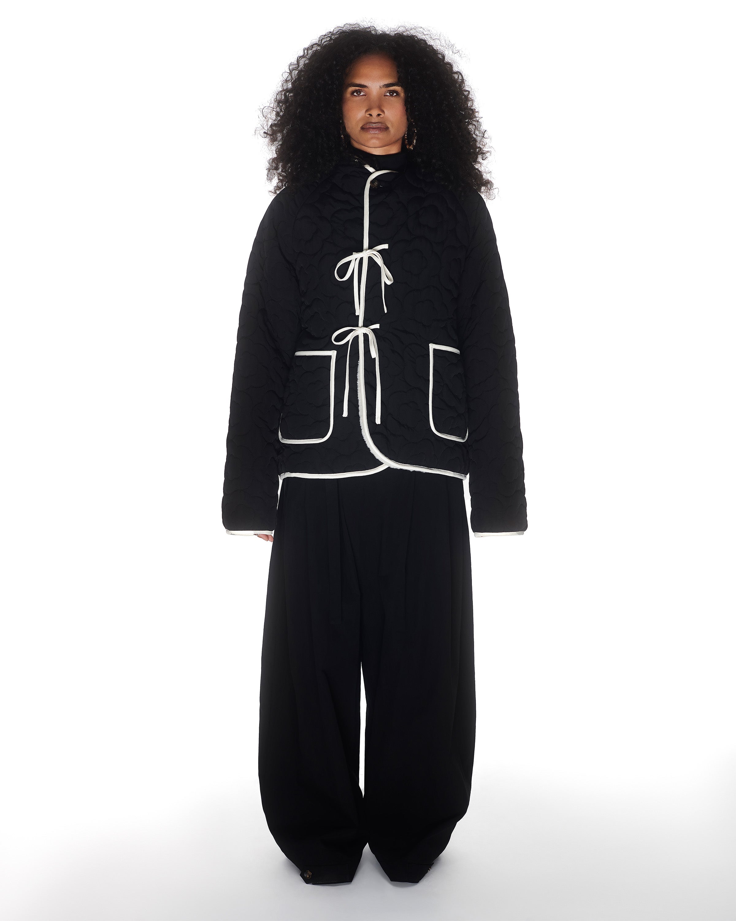 A person with long curly hair models Sundae School's BLACK IVORY REVERSIBLE FLOWER QUILTED JEOGORI FLEECE, a black Korean hanbok jacket with white trim and ties, paired with loose black pants against a white background.