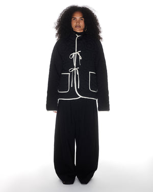 A person with long curly hair models Sundae School's BLACK IVORY REVERSIBLE FLOWER QUILTED JEOGORI FLEECE, a black Korean hanbok jacket with white trim and ties, paired with loose black pants against a white background.