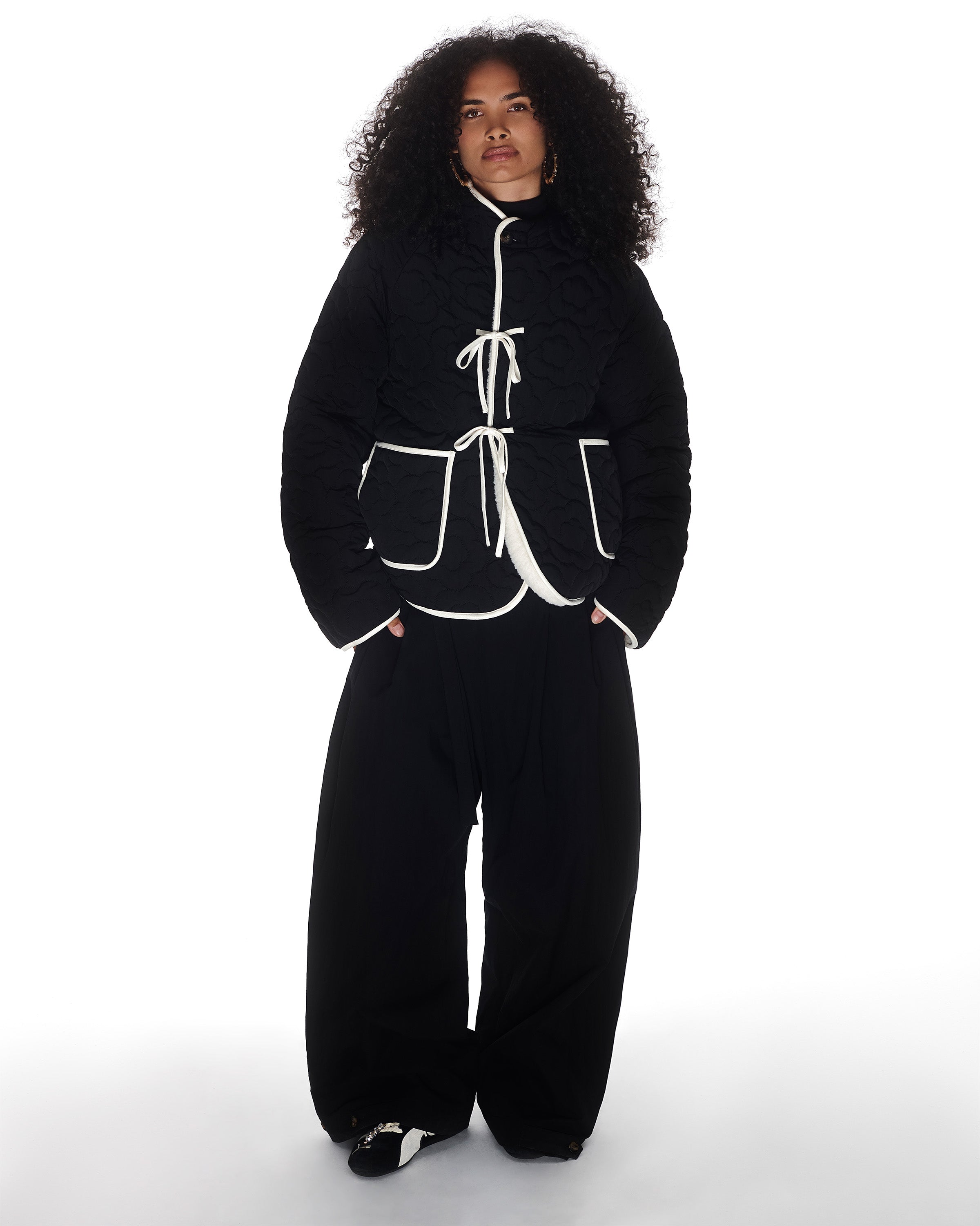 A person with long curly hair wears the Sundae School BLACK IVORY REVERSIBLE FLOWER QUILTED JEOGORI FLEECE, styled with wide-leg black pants and black shoes, standing against a plain white background.