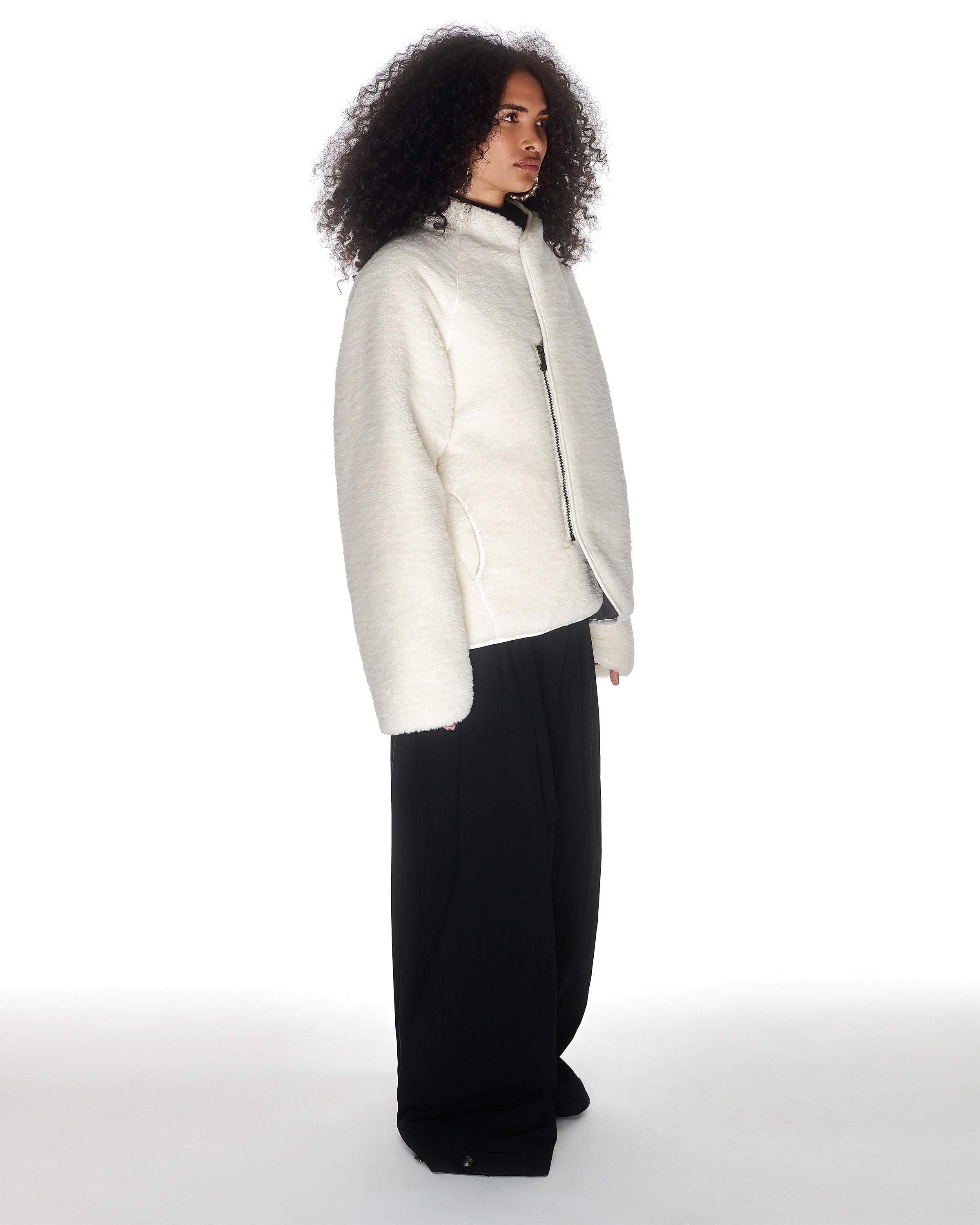 A person with curly hair stands sideways in Sundae School's BLACK IVORY REVERSIBLE FLOWER QUILTED JEOGORI FLEECE, featuring textured off-white sherpa and wide-leg black pants, set against a plain white background.
