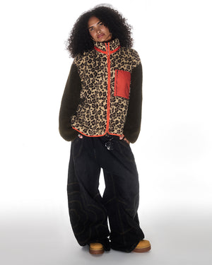 A person with curly hair wears the Sundae School Tiger Mom Fleece—leopard print with red trim and pocket—paired with baggy black jeans and tan boots, standing against a plain white background.
