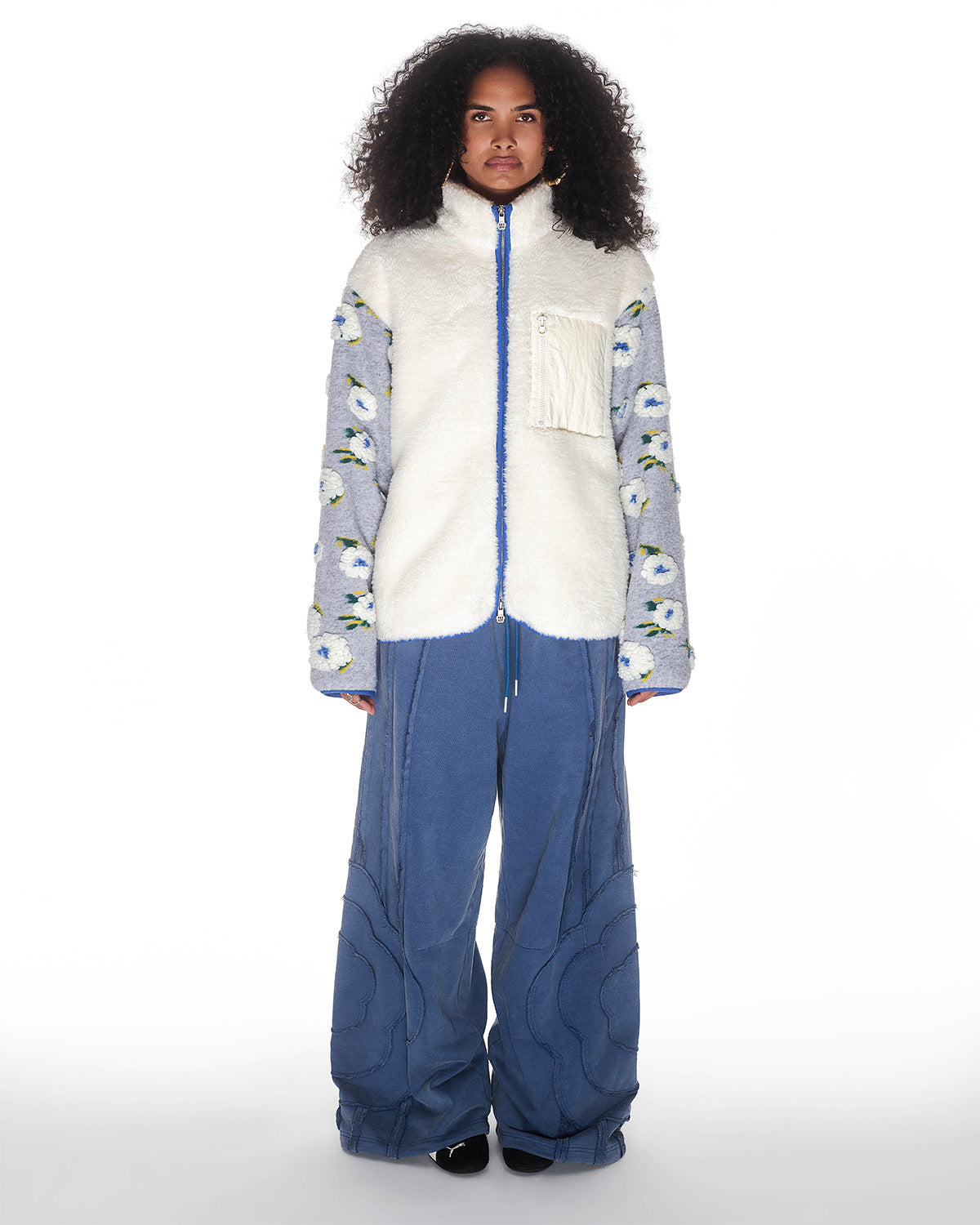 A woman with curly hair wears Sundae School's IVORY MUGUNGHWA FLEECE—a cream sherpa jacket with zip, pocket, and blue-and-yellow floral sleeves—paired with loose blue pants featuring curved seams, standing against a white background.