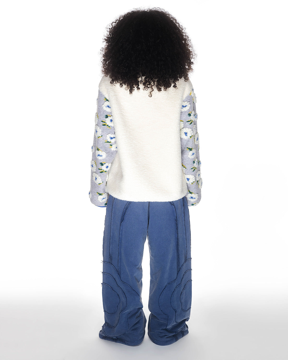 A person with curly hair faces away, wearing Sundae School's IVORY MUGUNGHWA FLEECE—white sherpa with patterned blue sleeves—and loose blue pants against a plain white background.