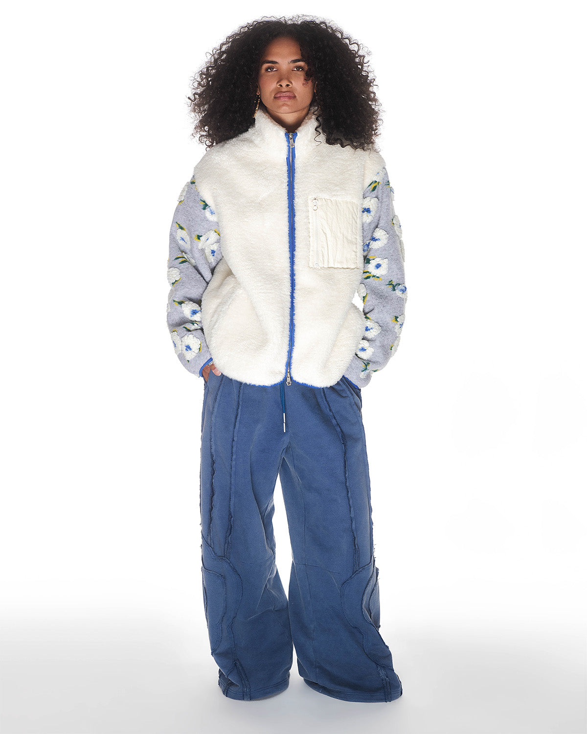 A person with curly hair faces forward, wearing Sundae School's IVORY MUGUNGHWA FLEECE vest over a blue floral hibiscus sweater and loose blue pants against a plain white background.