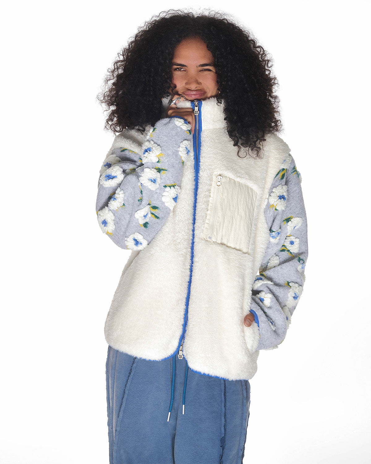 A person with curly hair smiles, wearing Sundae School's IVORY MUGUNGHWA FLEECE with white hibiscus sleeves and a white chest pocket, paired with blue pants against a white background.