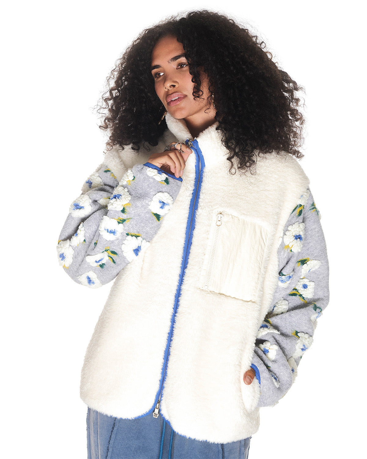A person with curly hair wears the Sundae School IVORY MUGUNGHWA FLEECE, a cream jacquard fleece jacket with a zipper, chest pocket, and grey sleeves featuring white flower patterns, set against a white background.