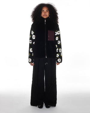 A person with curly hair faces forward, wearing Sundae School's Mugunghwa Fleece— a black zip-up with white hibiscus patterns on the sleeves and a maroon chest pocket—paired with wide-leg black pants and black shoes against a white background.