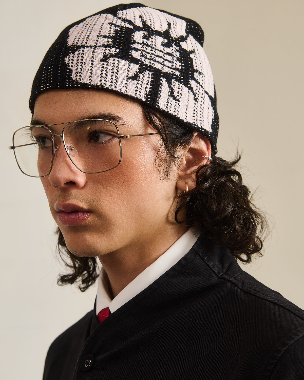 A person with long dark hair, large aviator glasses, a black jacket, white shirt with red tie, and the BLACK DDUL KNIT BEANIE by Sundae School looks to the side against a plain background.