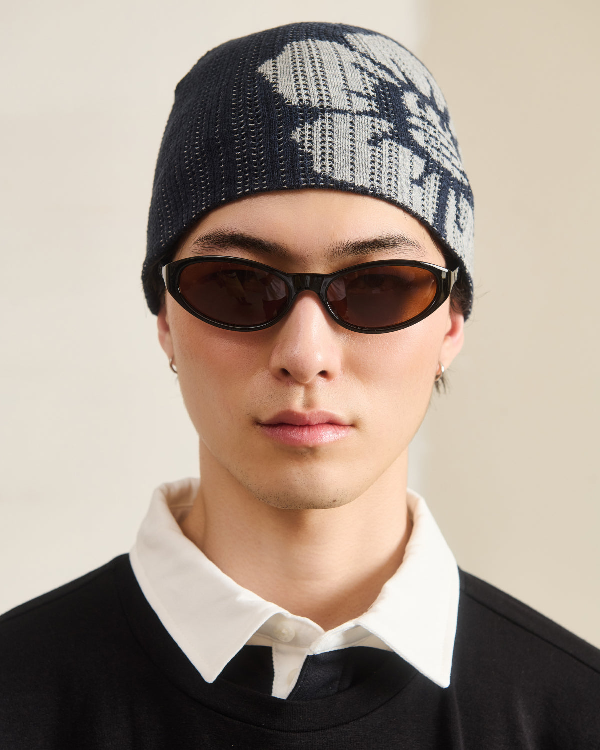 A young Asian man wears the Sundae School NAVY DDUL KNIT BEANIE, paired with brown oval sunglasses and a black crew neck shirt with a white collar, creating a minimalist look.