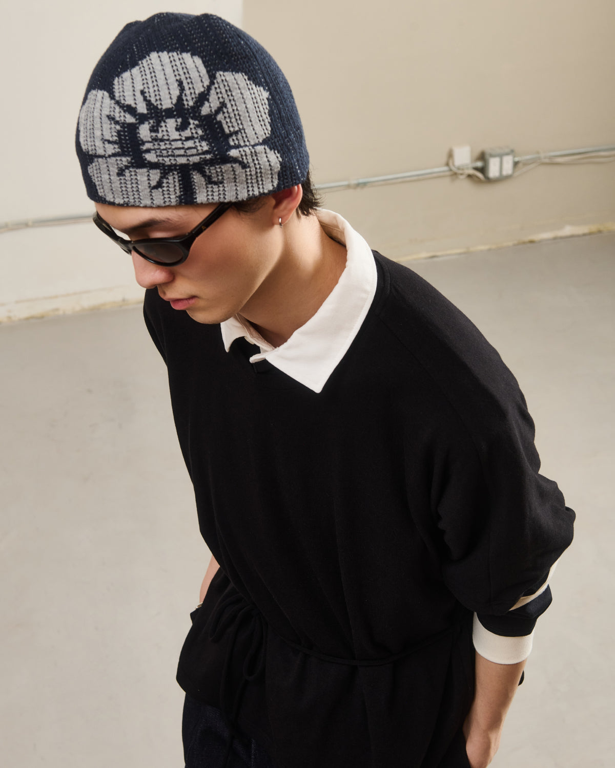 A person wearing black sunglasses, a Sundae School NAVY DDUL KNIT BEANIE, and a minimalist black top with a white collar stands indoors by a beige wall with exposed pipes.