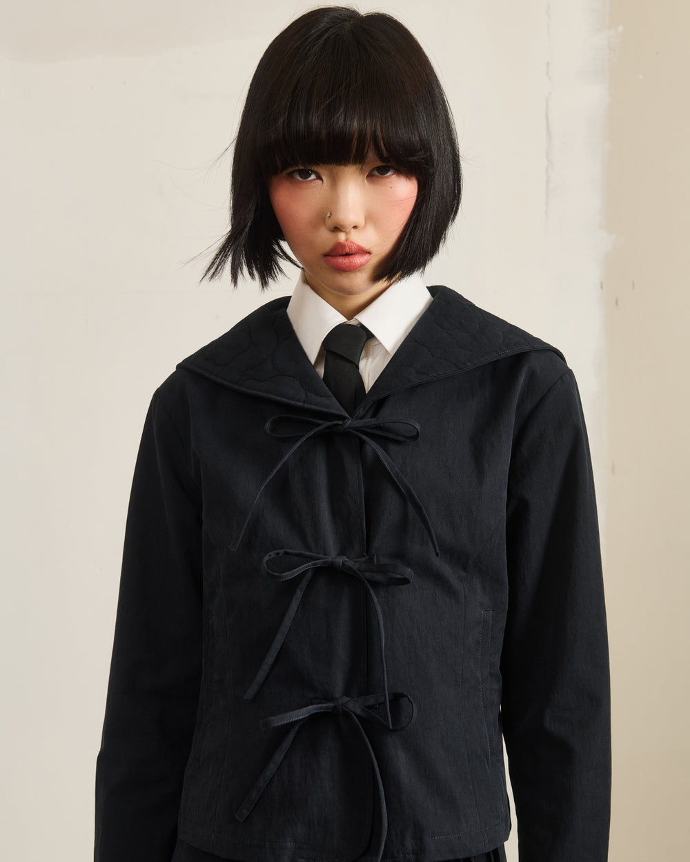 A person with short black hair and blunt bangs wears the Sundae School BLACK SAILOR COLLAR UNIFORM JACKET, featuring three bow ties and a wide collar over a white shirt and black tie, standing by a light-colored textured wall.