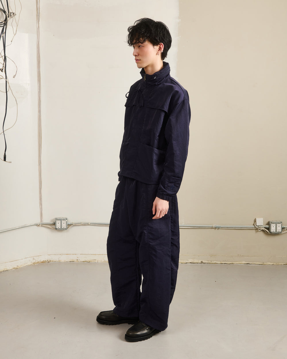 A person with short dark hair stands in a plain room wearing Sundae School's NAVY HANBOK BALLOON TRACK PANTS and black shoes. Exposed wires and outlets are visible on the beige wall behind them.