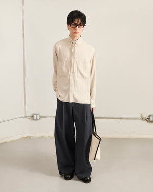 A person with short dark hair and glasses stands indoors, wearing Sundea School's IVORY HIGH NECK TIE SHIRT with oversized chest pockets, loose dark trousers, black shoes, and holding a beige tote bag against a minimal light background.