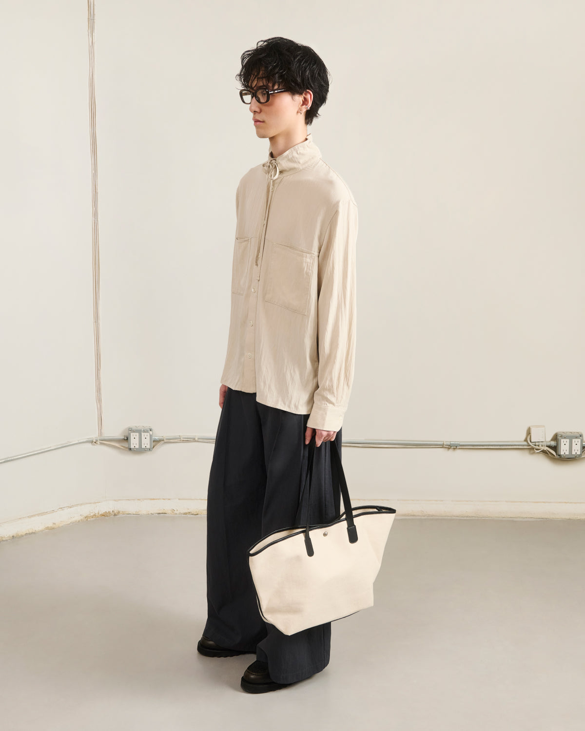 A person with short dark hair and glasses wears the Sundae School IVORY HIGH NECK TIE SHIRT, wide dark trousers, black shoes, and holds a cream tote bag with black handles while standing indoors.