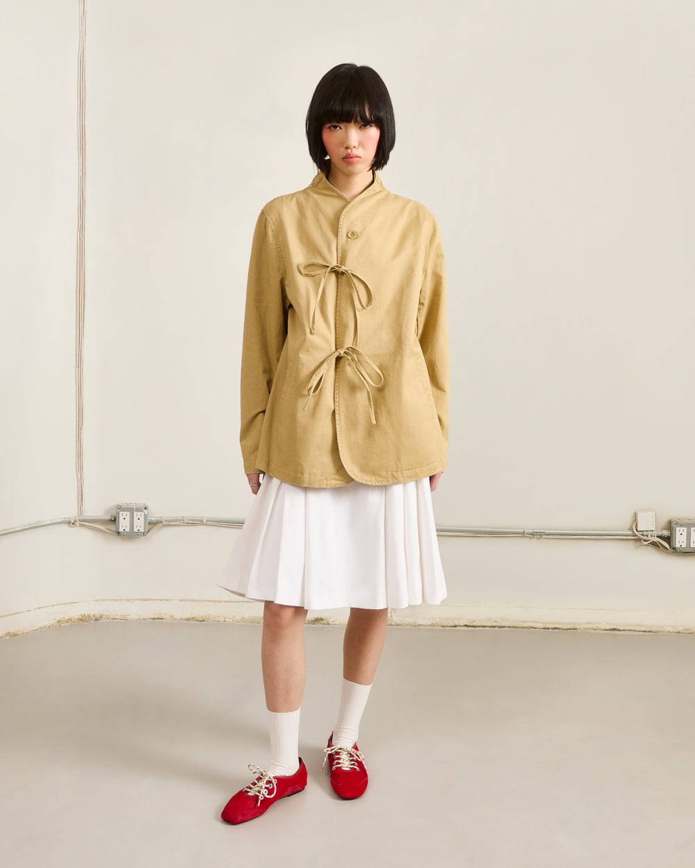 A person with a short black bob wears Sundae School’s TAN JEOGORI TIE JACKET, paired with a white pleated skirt, white socks, and red shoes, standing against a cream wall with exposed outlets and conduit.