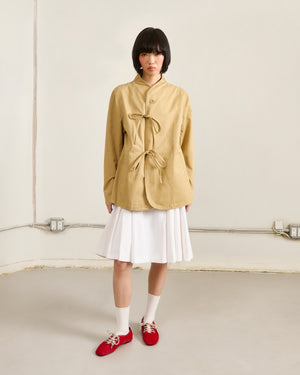 A person with a short black bob wears Sundae School’s TAN JEOGORI TIE JACKET, paired with a white pleated skirt, white socks, and red shoes, standing against a cream wall with exposed outlets and conduit.