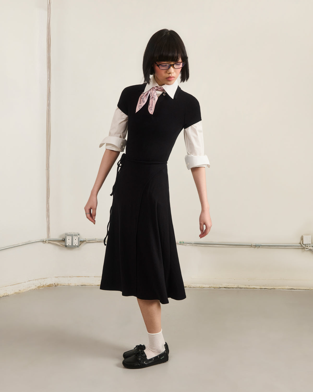 A person wearing the Sundae School CINDY POLO DRESS over a white shirt with rolled-up sleeves, styled with a pink neck scarf, white socks, and black shoes stands in a minimalist room with light walls and exposed outlets.