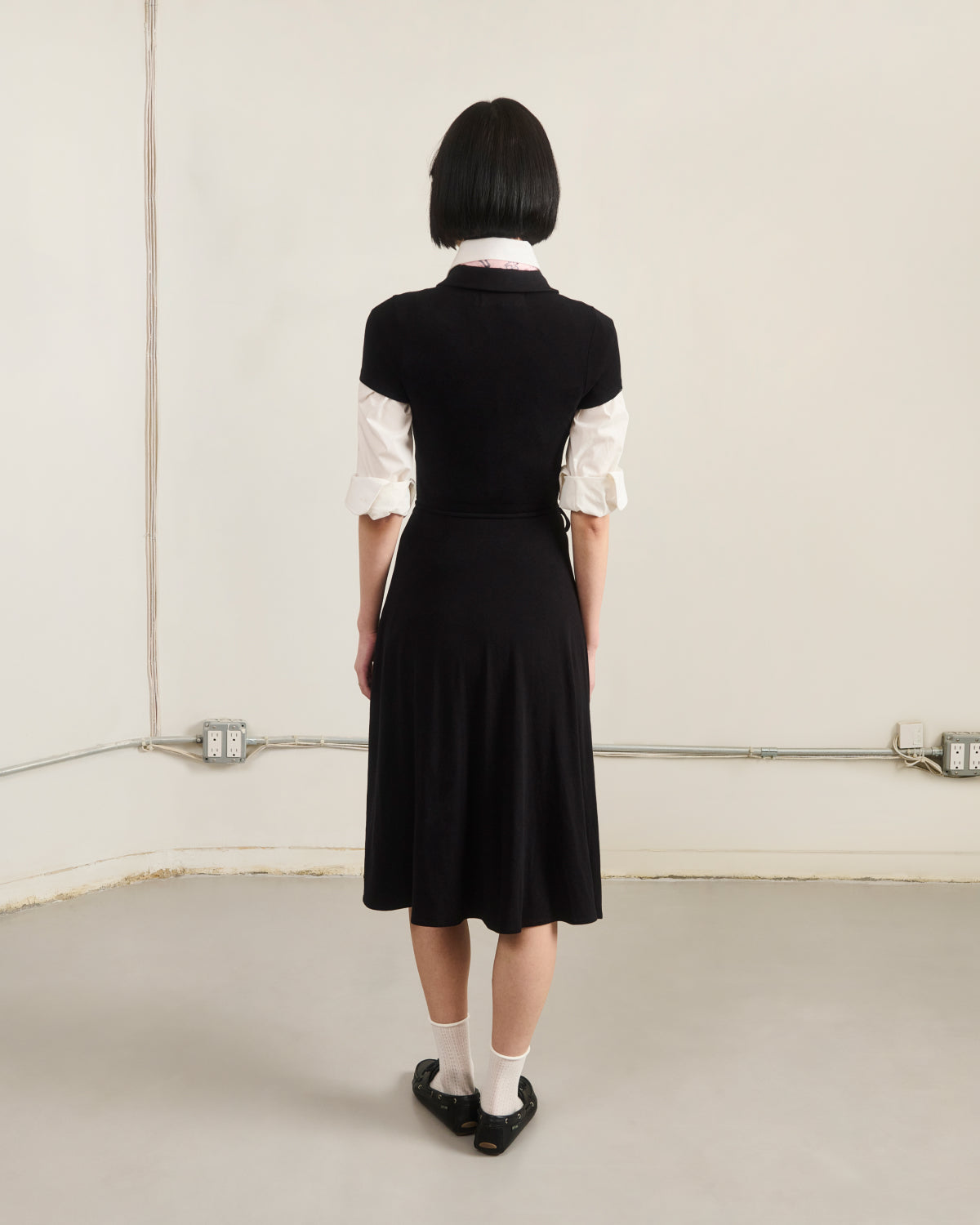 A person with short black hair faces a beige wall, wearing Sundae School's CINDY POLO DRESS over a white shirt, paired with white socks and black shoes. Exposed outlets and conduit lines are visible on the wall.