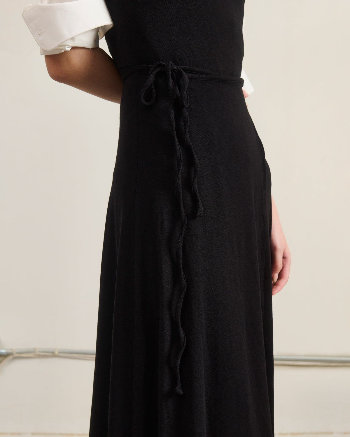 A person wears the Sundae School CINDY POLO DRESS, a black convertible wrap apron dress with a thin tied belt, over a white shirt with rolled-up sleeves, standing against a plain background.