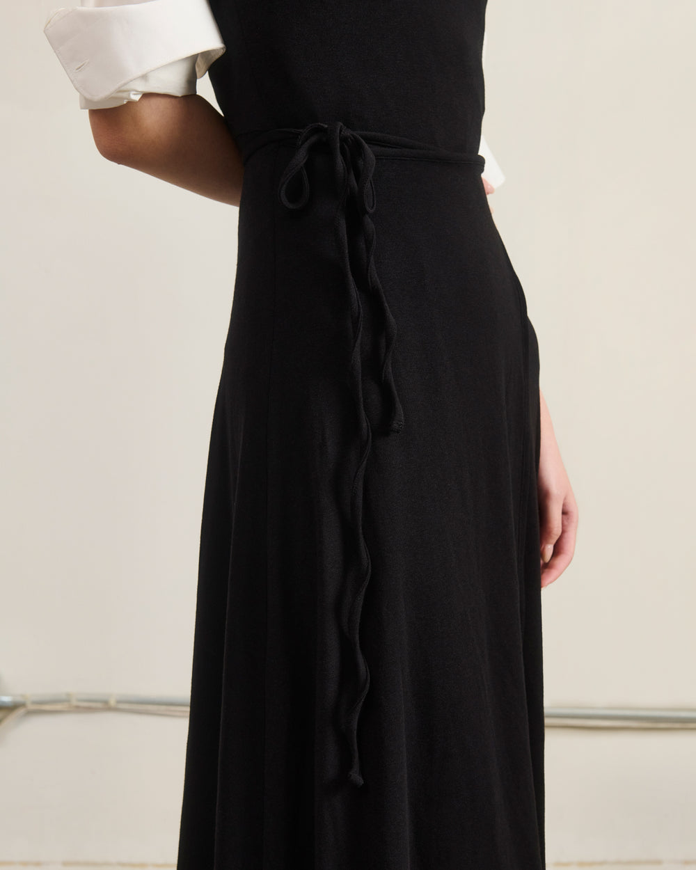 A person wearing the Sundae School BLACK CINDY POLO DRESS, styled over a white rolled-sleeve shirt and featuring a thin waist tie, is shown from the torso up against a plain background.