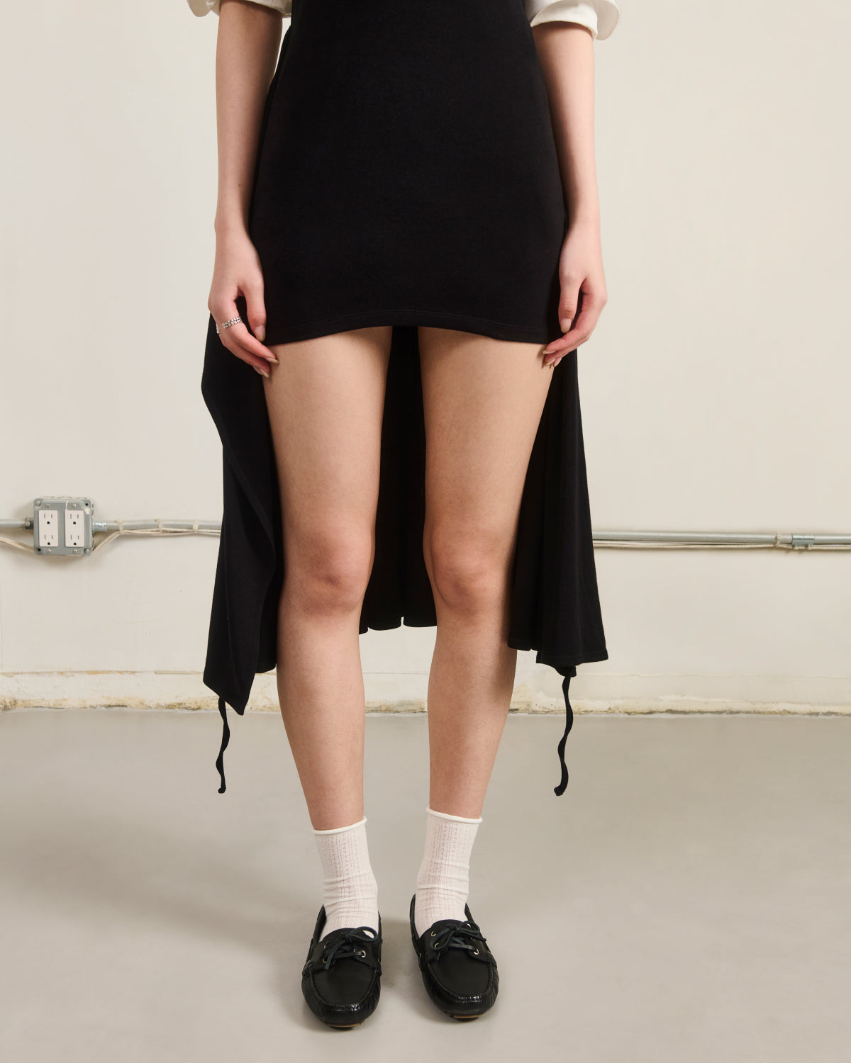 Indoors against a plain wall, a person wears the Sundae School CINDY POLO DRESS—a fitted, black midi dress with a high-low hem—paired with white socks and black loafers, hands resting at their sides.