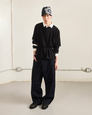 A person stands indoors against a plain wall, wearing Sundae School's BLACK BAERAE T-SHIRT over a white collared shirt, a black beanie with a light pattern, wide dark pants, and black shoes. Their hands are in their pockets.