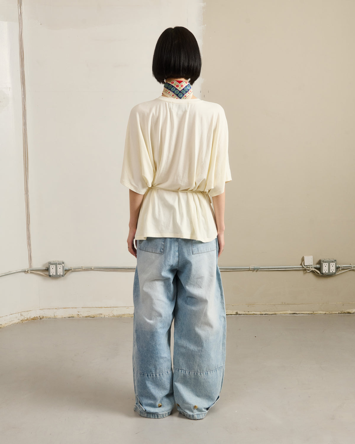 A person with short black hair stands facing a beige wall, wearing Sundae School’s IVORY BAERAE T-SHIRT, wide light blue jeans, and a patterned scarf. Exposed outlets and electrical wiring are visible in the room.