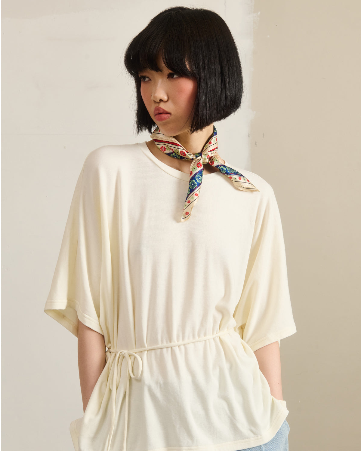 A person with a short black bob wears the Sundae School IVORY BAERAE T-SHIRT, which has a loose fit and gathered waist, with a colorful scarf around their neck, standing against a plain light background.